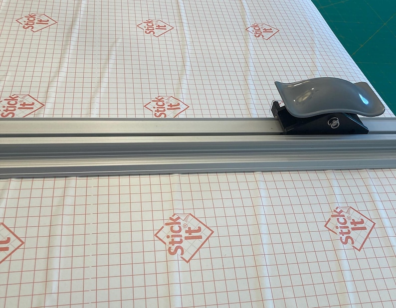 May include: A silver cutting mat with a grid pattern and the words "Stick It" printed on it. A gray plastic cutter is on the mat.