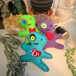 May include: Three handmade felt voodoo dolls in green, purple, and blue. Each doll has button eyes, stitched mouths, and decorative accents like hearts and bows. The dolls are arranged on a white surface.