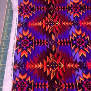 Puede incluir: Una tela vibrante con un patr&oacute;n geom&eacute;trico en tonos de morado, rojo, naranja y marr&oacute;n. El dise&ntilde;o presenta formas angulares repetidas y motivos de diamantes. La tela est&aacute; etiquetada como "Timeless Treasures Fabrics of SoHo, LLC."