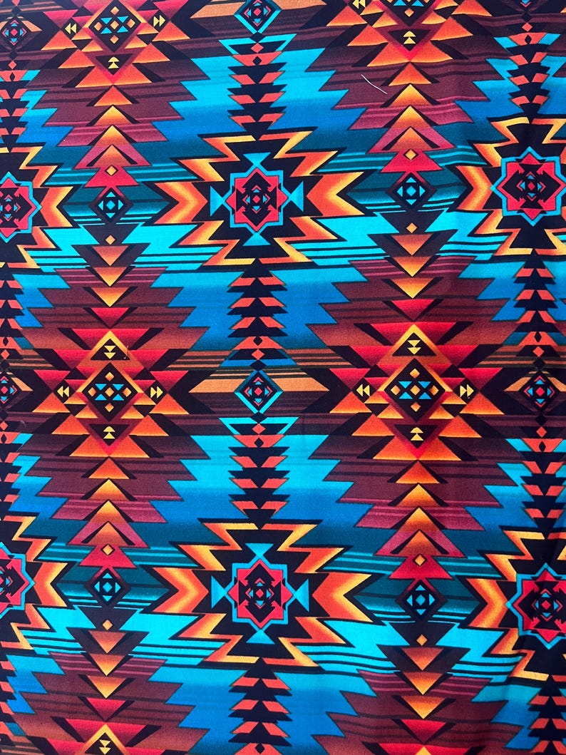 Op de afbeelding: Een levendige stof met een geometrisch patroon in tinten turquoise, oranje, rood en bruin. Het ontwerp bevat zigzags, ruitvormen en pijlvormige vormen, wat een gedurfde, door het zuidwesten ge&iuml;nspireerde esthetiek cre&euml;ert.