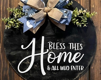 May include: A black round wooden sign with the text "Bless This Home & All Who Enter" in white cursive lettering. The sign is decorated with a blue and brown bow and green foliage.