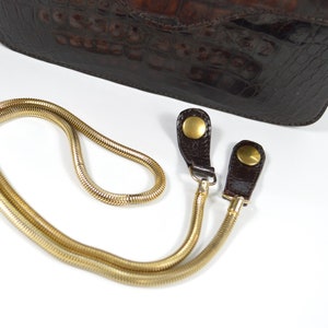 May include: A gold chain strap with two brown leather loops and gold metal clasps. The strap is designed to be used with a handbag.