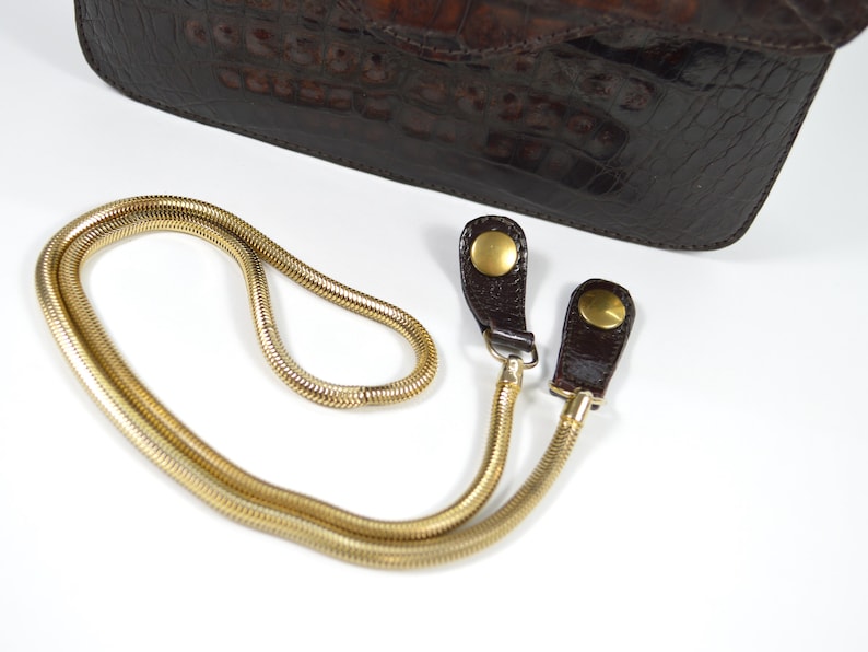 May include: A gold chain strap with two brown leather loops and gold metal clasps. The strap is designed to be used with a handbag.