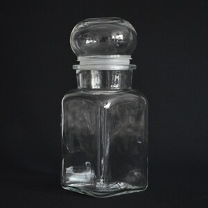 May include: A clear glass jar with a square body and a round glass lid. The jar has a smooth, polished surface.