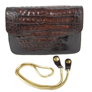 May include: A brown crocodile leather clutch purse with a gold chain strap. The purse has a flap closure and a textured crocodile pattern.