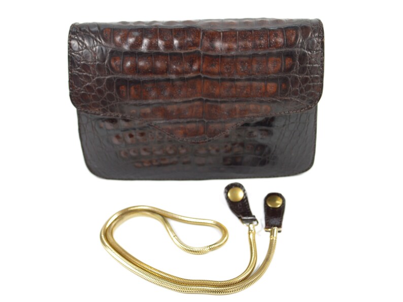 May include: A brown crocodile leather clutch purse with a gold chain strap. The purse has a flap closure and a textured crocodile pattern.