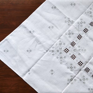 Puede incluir: Mantel de mesa de lino blanco con un delicado diseño bordado en hilo gris. El diseño presenta un patrón repetitivo de flores estilizadas y formas geométricas.