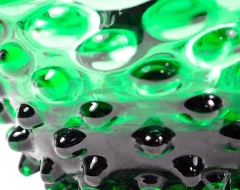 Op de afbeelding: Een levendig smaragdgroen glazen object met een getextureerd oppervlak met talloze verhoogde, afgeronde bultjes en inkepingen. Het doorschijnende materiaal vangt het licht, waardoor verschillende tinten groen ontstaan op de tactiele, gedimde buitenkant.