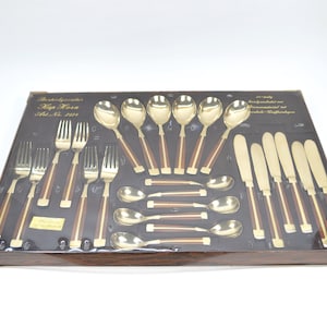 Op de afbeelding: Een set gouden bestek met houten handvatten in een houten doos. De doos heeft een label met de tekst "Besteckgarnitur Kip Torn Art. Nr. 201".