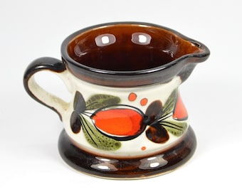May include: A ceramic creamer with a brown interior and rim, a white body, and a brown base and handle. The body is decorated with orange, green, and brown floral designs. The creamer has a spout for easy pouring.
