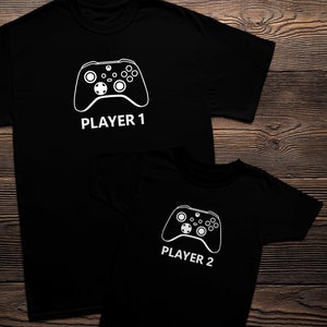 May include: Two black t-shirts with white text and a white graphic of a video game controller. The larger shirt says "Player 1" and the smaller shirt says "Player 2".