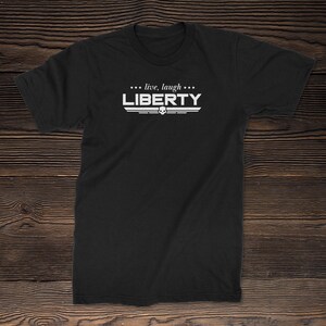 Può includere: T-shirt nera con la scritta "LIBERTY" in bianco, sopra un disegno di teschio e ali. La frase "... live, laugh ..." è sopra la parola "LIBERTY". La maglietta è realizzata in un materiale morbido.