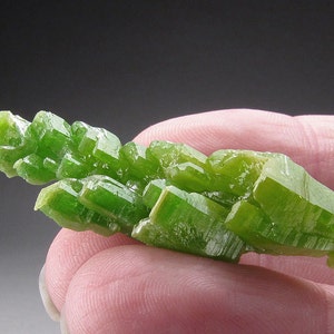 Puede incluir: Un grupo de cristales alargados translúcidos de color verde brillante con estrías. Los cristales están dispuestos en forma de abanico.