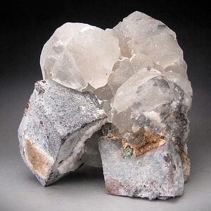 Puede incluir: Un grupo de cristales de calcita transparentes y translúcidos sobre una matriz de roca gris y marrón. Los cristales están agrupados en una forma redondeada, y algunos tienen una apariencia ligeramente lechosa. La matriz es rugosa y texturizada.