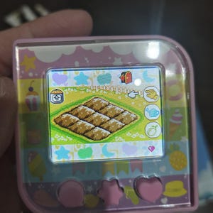 May include: A pink handheld digital pet device with a square screen displaying a pixelated grid of brown food items. The device features a colorful border with icons of stars, hearts, and fruit, and three pink buttons at the bottom in circular and star shapes.