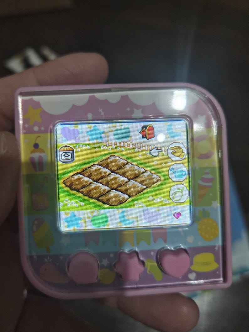 May include: A pink handheld digital pet device with a square screen displaying a pixelated grid of brown food items. The device features a colorful border with icons of stars, hearts, and fruit, and three pink buttons at the bottom in circular and star shapes.