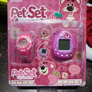 May include: Pink and purple toy set with a pink watch, a keychain with a pink bear, and a purple handheld game with a pink bear on the screen. The packaging is pink with a strawberry design and the text "Pet Set Exquisite Pet Set".
