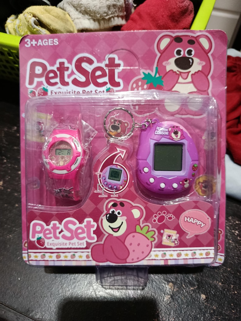 May include: Pink and purple toy set with a pink watch, a keychain with a pink bear, and a purple handheld game with a pink bear on the screen. The packaging is pink with a strawberry design and the text "Pet Set Exquisite Pet Set".