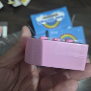 May include: A rectangular light pink plastic electronic device featuring three small pink buttons along the top edge. The device is positioned in front of blue packaging that displays a graphic of a colorful rainbow and white clouds.