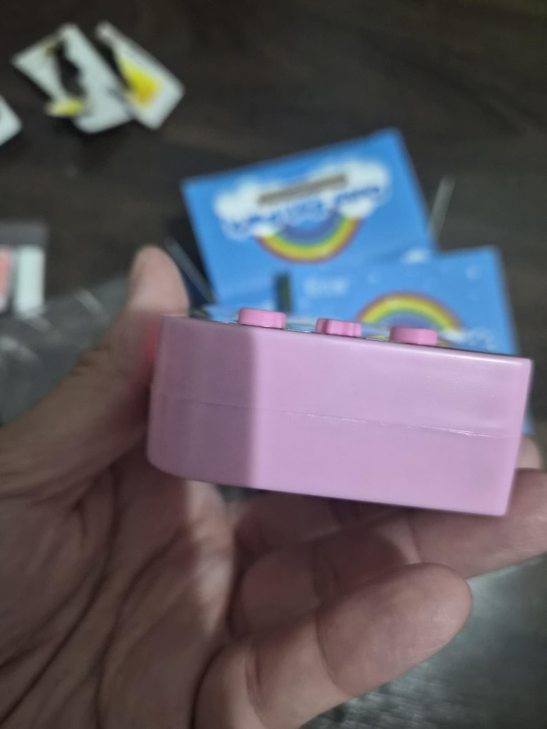 May include: A rectangular light pink plastic electronic device featuring three small pink buttons along the top edge. The device is positioned in front of blue packaging that displays a graphic of a colorful rainbow and white clouds.