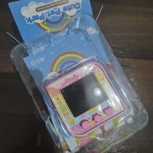 May include: A pink handheld electronic pet game console with a square screen and three shaped buttons, including a circle, star, and heart. The device is encased in clear plastic packaging with a blue backing card featuring a rainbow and the text Cute Pet Park.