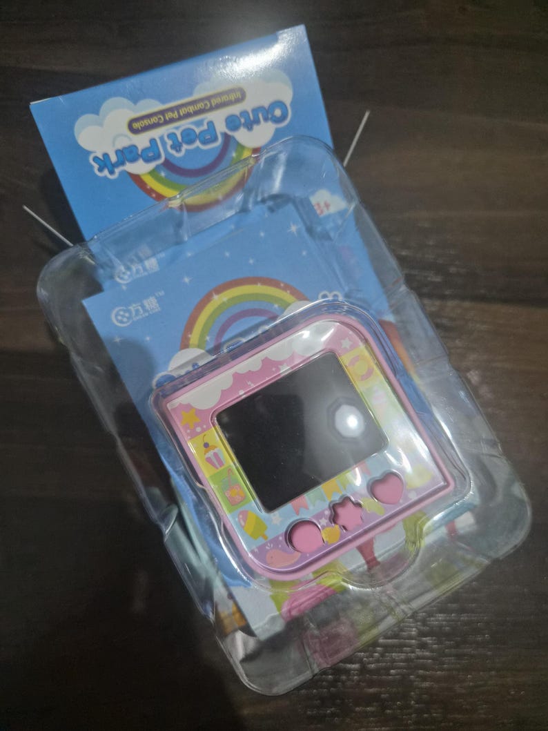 May include: A pink handheld electronic pet game console with a square screen and three shaped buttons, including a circle, star, and heart. The device is encased in clear plastic packaging with a blue backing card featuring a rainbow and the text Cute Pet Park.