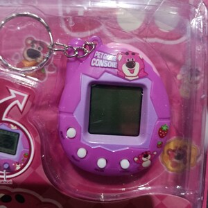 May include: A purple and pink keychain pet game console with a cartoon bear on the front. The console has a screen, buttons, and a strawberry graphic. The text "PET GAME CONSOLE" is printed on the front.
