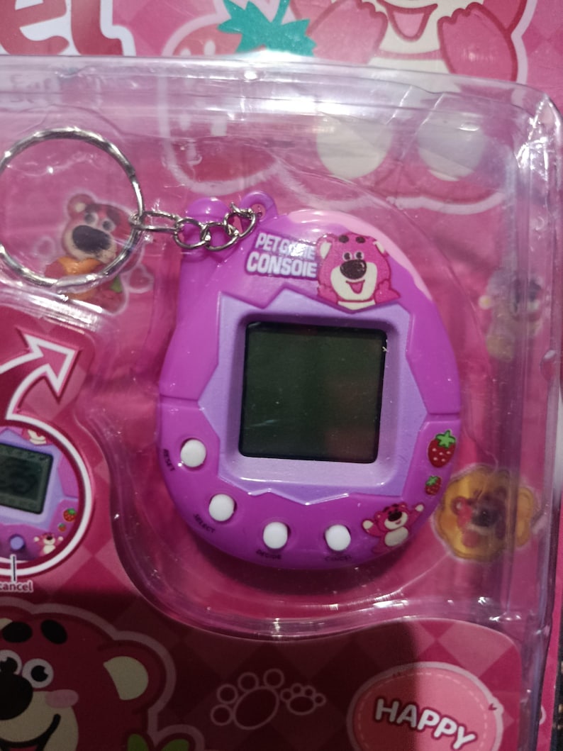 May include: A purple and pink keychain pet game console with a cartoon bear on the front. The console has a screen, buttons, and a strawberry graphic. The text "PET GAME CONSOLE" is printed on the front.