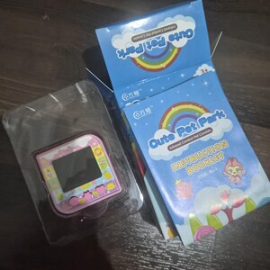 May include: A pink handheld digital pet console with a square screen and buttons, housed in a clear plastic tray next to its blue cardboard packaging. The box features a rainbow graphic, the text Cute Pet Park, and an instruction booklet.
