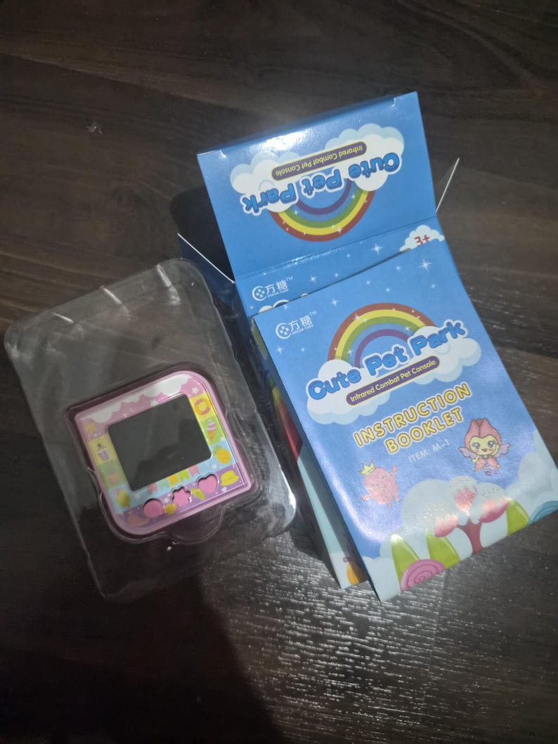 May include: A pink handheld digital pet console with a square screen and buttons, housed in a clear plastic tray next to its blue cardboard packaging. The box features a rainbow graphic, the text Cute Pet Park, and an instruction booklet.