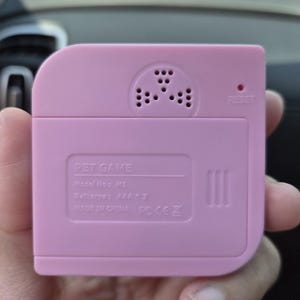 May include: A square, light pink plastic pet game device with rounded corners. The back features a speaker grille, a small reset hole, and molded text reading PET GAME, Model No: M1, Batterys: AAA * 3, MADE IN CHINA, and regulatory symbols.