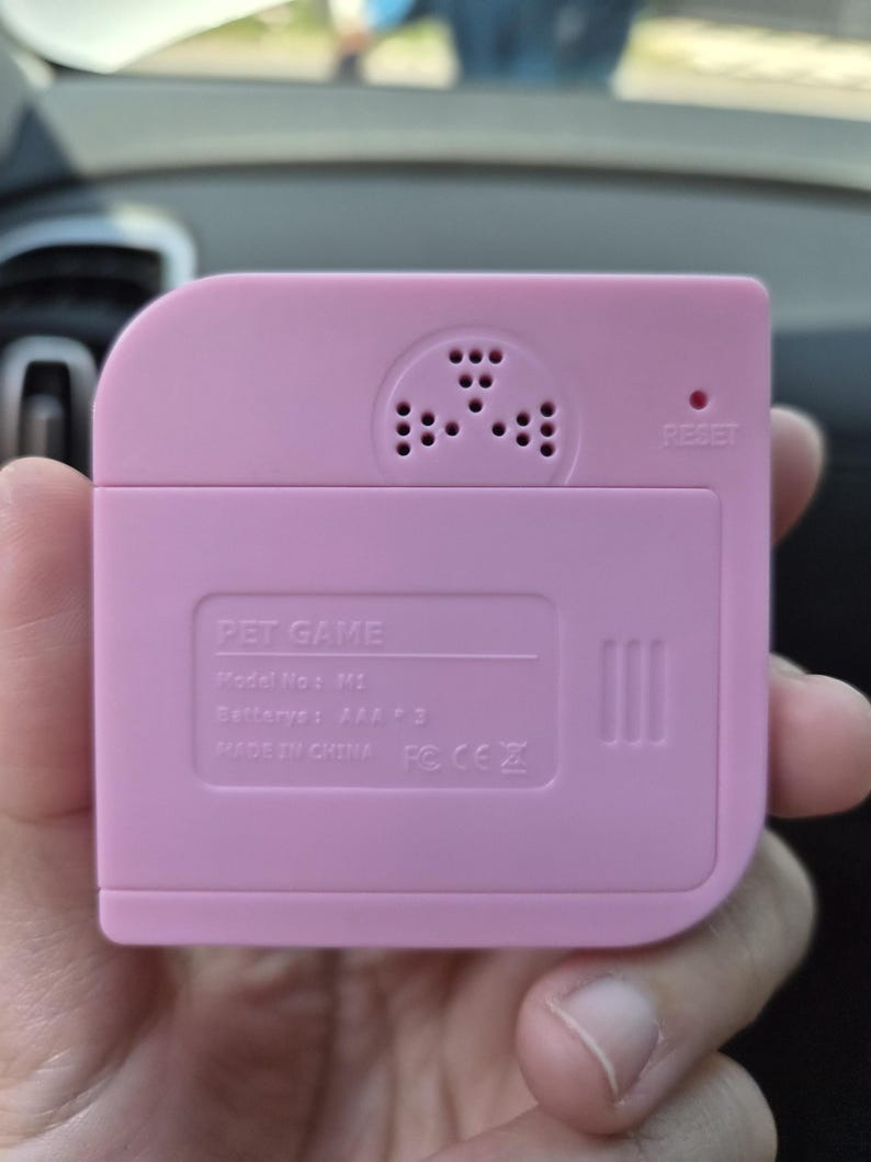 May include: A square, light pink plastic pet game device with rounded corners. The back features a speaker grille, a small reset hole, and molded text reading PET GAME, Model No: M1, Batterys: AAA * 3, MADE IN CHINA, and regulatory symbols.