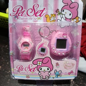 May include: Pink packaging with a pink Hello Kitty character holding a flower. The packaging contains a pink watch and a pink keychain with a Hello Kitty character on it. The packaging says "Pet Set Exquisite Pet Set" and "Sweet!"