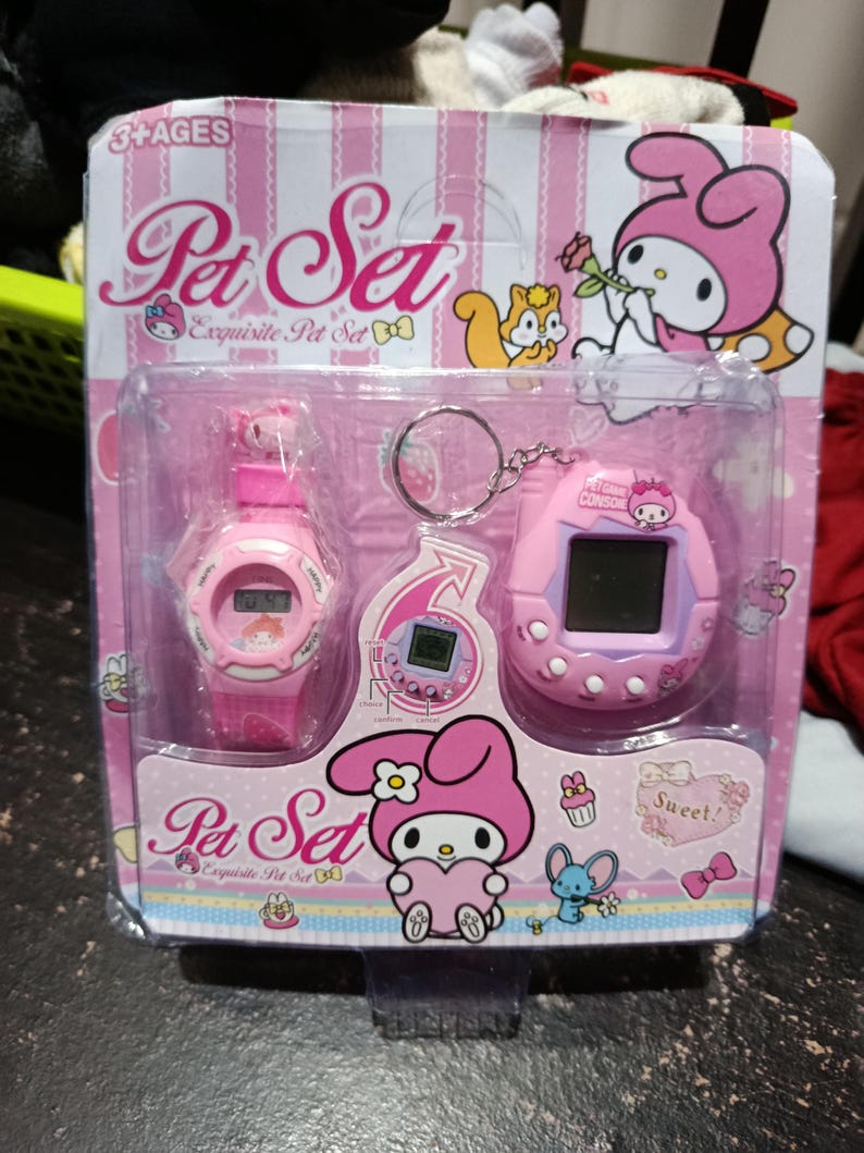May include: Pink packaging with a pink Hello Kitty character holding a flower. The packaging contains a pink watch and a pink keychain with a Hello Kitty character on it. The packaging says "Pet Set Exquisite Pet Set" and "Sweet!"