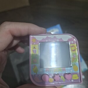 May include: A square, light pink digital pet device featuring a central screen surrounded by a colorful border with icons of ice cream, pineapples, and stars. Below the screen are three pink buttons shaped like a circle, a star, and a heart.