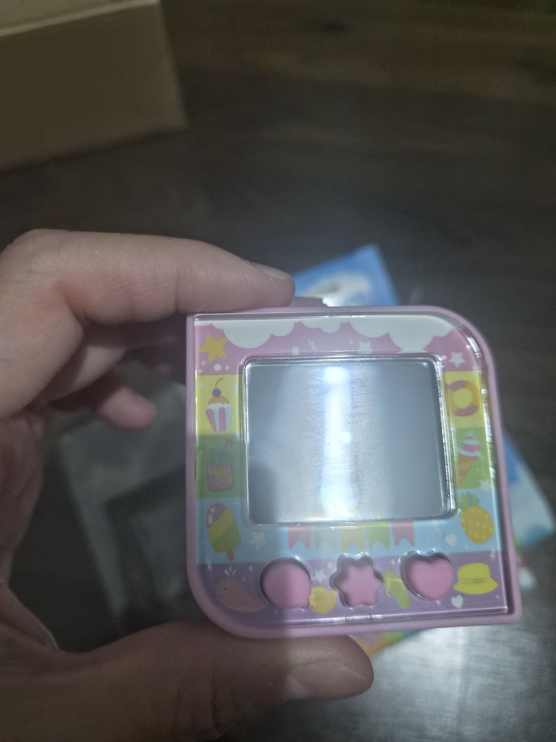 May include: A square, light pink digital pet device featuring a central screen surrounded by a colorful border with icons of ice cream, pineapples, and stars. Below the screen are three pink buttons shaped like a circle, a star, and a heart.