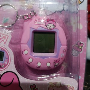 May include: Pink Hello Kitty keychain pet game console with a digital screen, buttons, and a Hello Kitty character on the front. The console is in a plastic packaging with a pink background and Hello Kitty graphics.