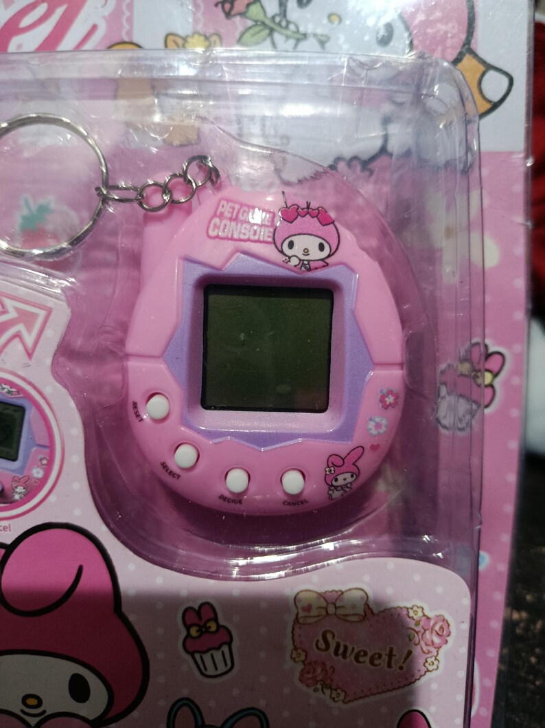 May include: Pink Hello Kitty keychain pet game console with a digital screen, buttons, and a Hello Kitty character on the front. The console is in a plastic packaging with a pink background and Hello Kitty graphics.