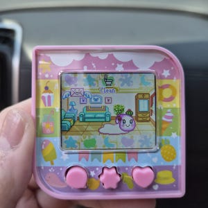 May include: A pink digital pet device with a square screen displaying a pixelated character in a room. The device frame features colorful illustrations of sweets, fruits, and stars. Three pink buttons are located below the screen, including a central star-shaped button flanked by two circular and heart-shaped buttons.