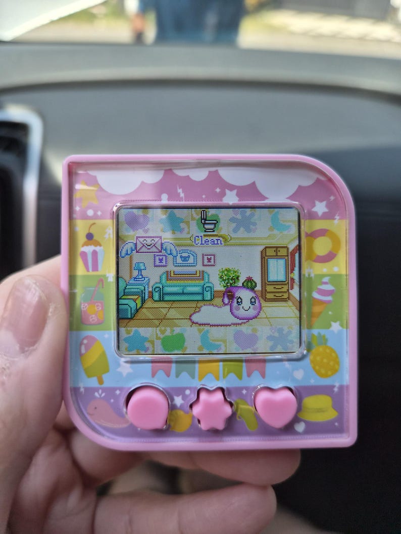 May include: A pink digital pet device with a square screen displaying a pixelated character in a room. The device frame features colorful illustrations of sweets, fruits, and stars. Three pink buttons are located below the screen, including a central star-shaped button flanked by two circular and heart-shaped buttons.