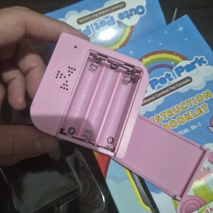 May include: A light pink plastic electronic game console with an open battery compartment showing three slots for AAA batteries and a speaker grille. The console is positioned over a blue instruction booklet labeled Pet Park Infrared Combat Pet Console.