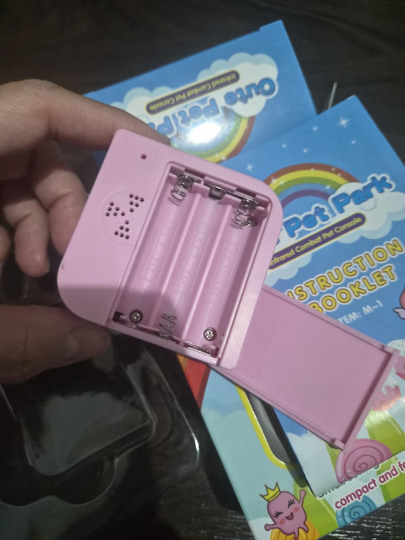 May include: A light pink plastic electronic game console with an open battery compartment showing three slots for AAA batteries and a speaker grille. The console is positioned over a blue instruction booklet labeled Pet Park Infrared Combat Pet Console.
