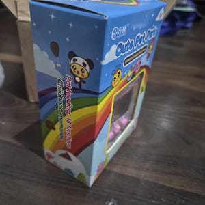 May include: A rectangular blue cardboard box for a digital pet toy featuring a rainbow graphic, cartoon characters, and the text Cute Pet Park, Pet feeding, 18 functions, and Childhood memories. The packaging includes a clear plastic window.