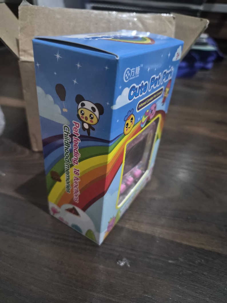 May include: A rectangular blue cardboard box for a digital pet toy featuring a rainbow graphic, cartoon characters, and the text Cute Pet Park, Pet feeding, 18 functions, and Childhood memories. The packaging includes a clear plastic window.