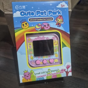 May include: A pink handheld electronic pet console in a blue box labeled Cute Pet Park and Infrared Combat Pet Console. The packaging features a rainbow, cartoon characters, and the text Small soft light display, compact and fashionable!