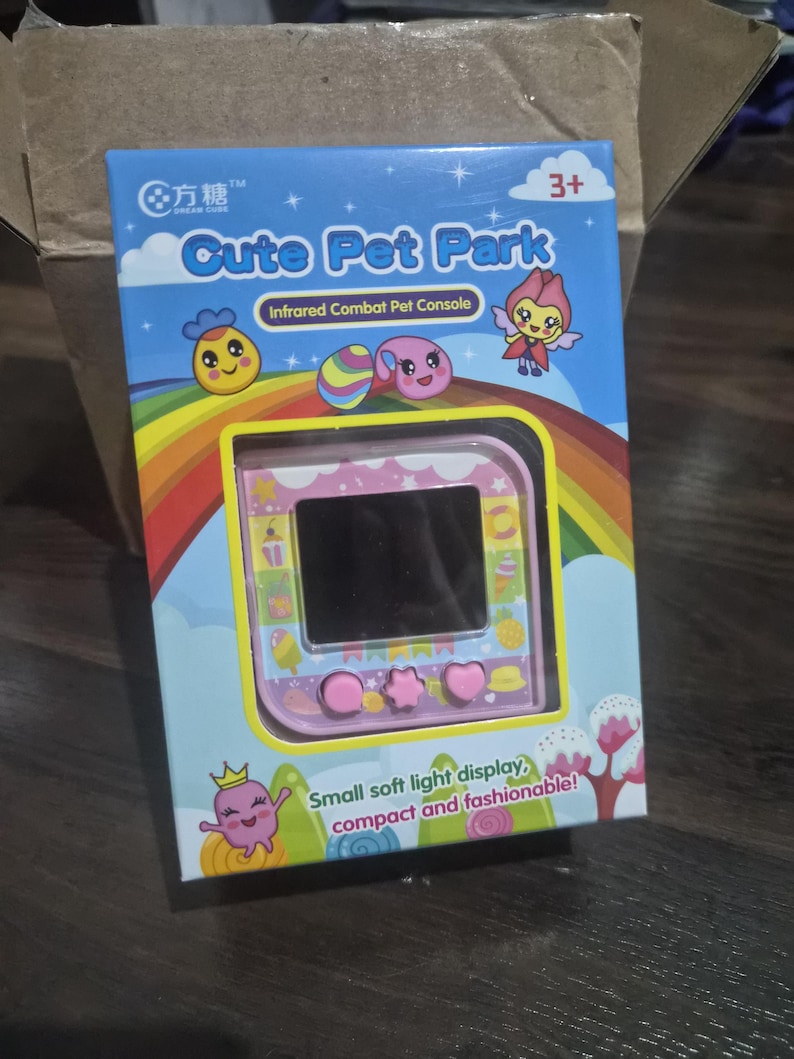 May include: A pink handheld electronic pet console in a blue box labeled Cute Pet Park and Infrared Combat Pet Console. The packaging features a rainbow, cartoon characters, and the text Small soft light display, compact and fashionable!