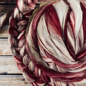 May include: Close-up of a skein of hand-dyed yarn in shades of burgundy, white, and gold. The yarn is spun and plied, with a braided section visible. The fibers have a soft, textured appearance, suitable for knitting or other fiber arts projects.