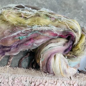 Puede incluir: Primer plano de una madeja de hilo colorida. El hilo presenta una mezcla de colores, incluyendo tonos de morado, rosa, verde y blanco. El hilo tiene un aspecto suave y texturizado con hebras y fibras visibles, adecuado para proyectos de arte textil.
