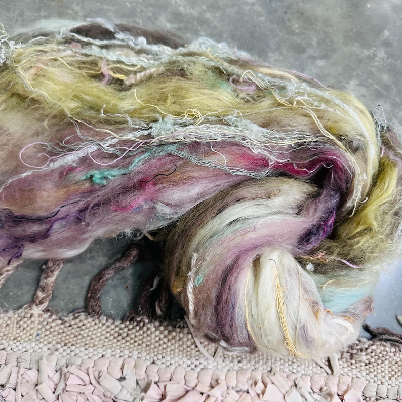 Puede incluir: Primer plano de una madeja de hilo colorida. El hilo presenta una mezcla de colores, incluyendo tonos de morado, rosa, verde y blanco. El hilo tiene un aspecto suave y texturizado con hebras y fibras visibles, adecuado para proyectos de arte textil.