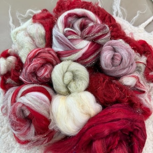 May include: A collection of colorful fiber art materials, including red, white, and pink wool roving. Some of the wool has a metallic silver thread woven in. The fibers are arranged in a variety of shapes, including loose piles and tightly wound rolls.